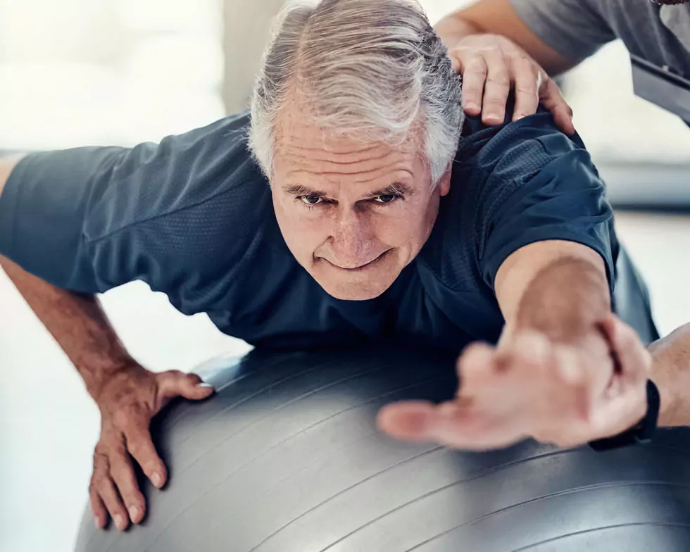 Gleichgewichtsübung älterer Herr bei Gleichgewichtsübung mit einem Gymnastikball. Physiotherapeut unterstützt und hilft dem Patienten