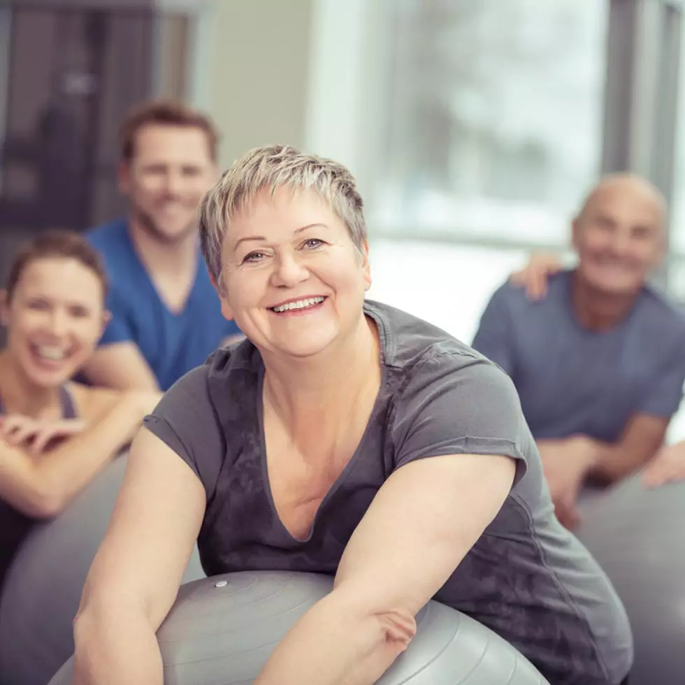 Gruppe beim Sport Lächelnde ältere Frau, die einen Pilates-Kurs im Fitnessstudio genießt und sich auf ihren Ball stützt und in die Kamera lächelt, während die Klasse hinter ihr steht