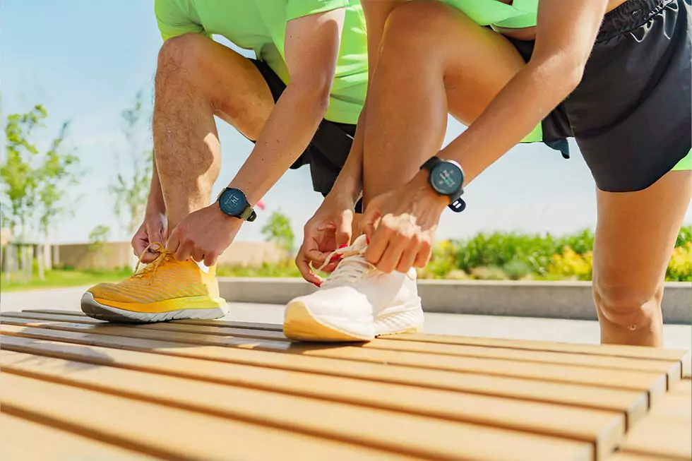 Zwei Sportler binden ihre Schnürsenkel vor einem morgendlichen Lauf in einem Park umgeben von Grün und klarem blauen Himmel